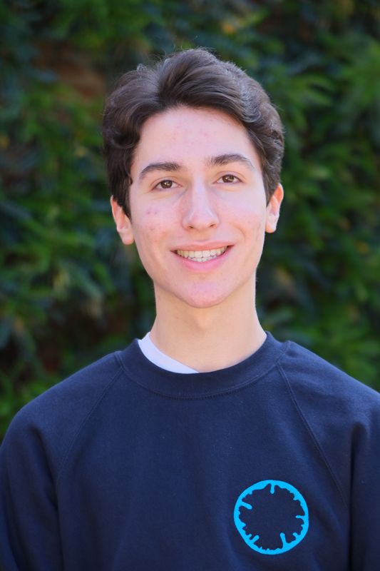 Portrait photograph of Joseph Vasconcelos Reed smiling in front of a leafy background