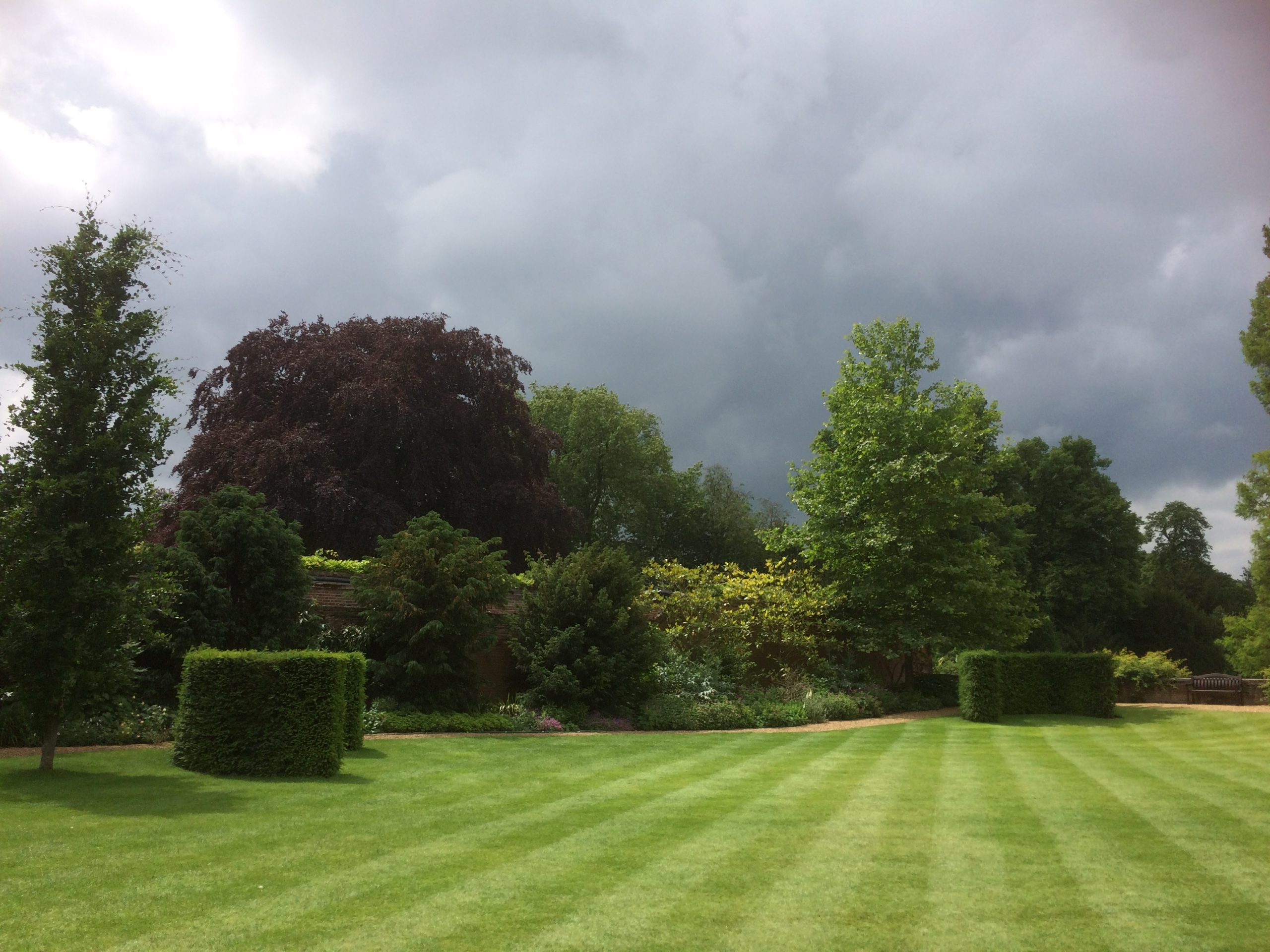 View down the Fellows' Garden towards the river. The lawn is green and striped and storm clouds are gathering overhead.