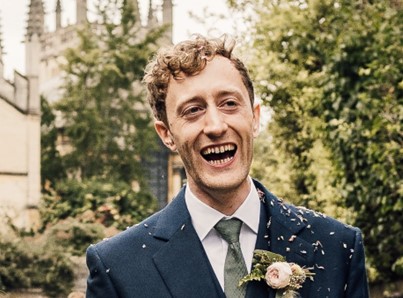 Tom Leece dressed in a navy suit and pink corsage, smiling towards the camera