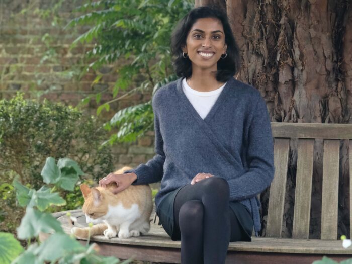 Smiling woman (Caitlin Rajan) stroking Silly the cat in Trinity Hall while sitting under a tree.