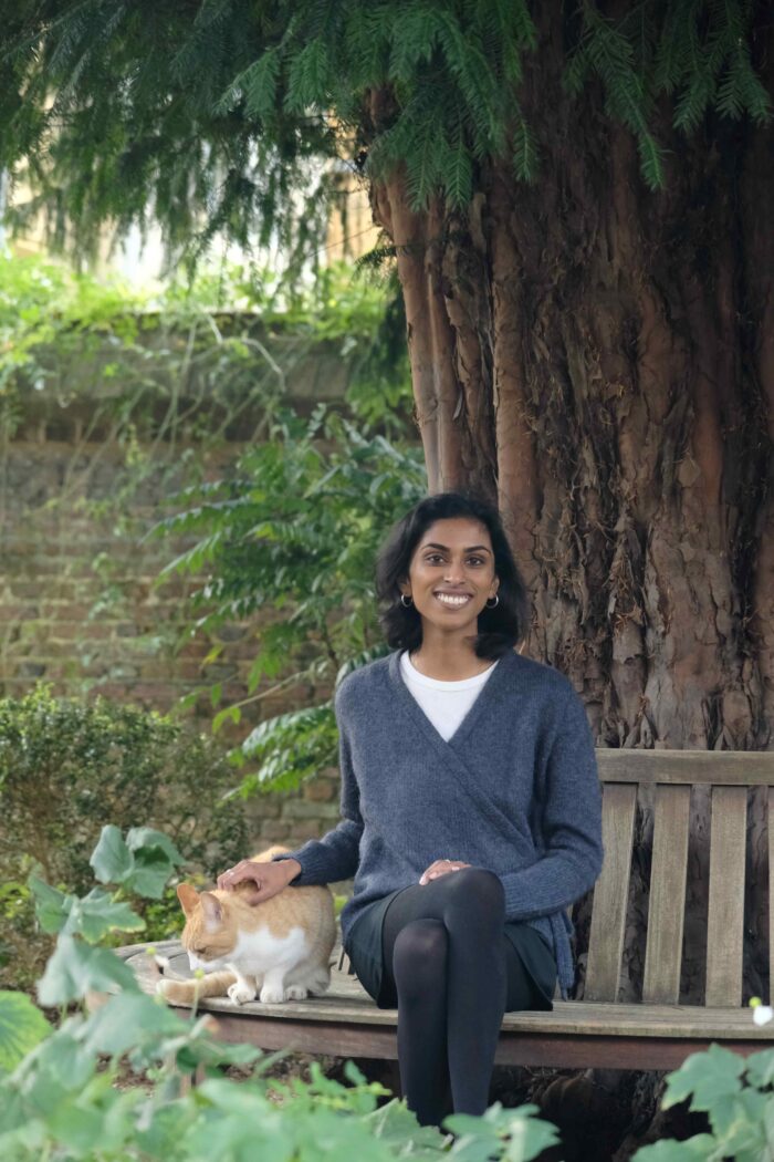Caitlin Rajan is smiling as she strokes Silly the cat. They are both under a tree in a garden at Trinity Hal