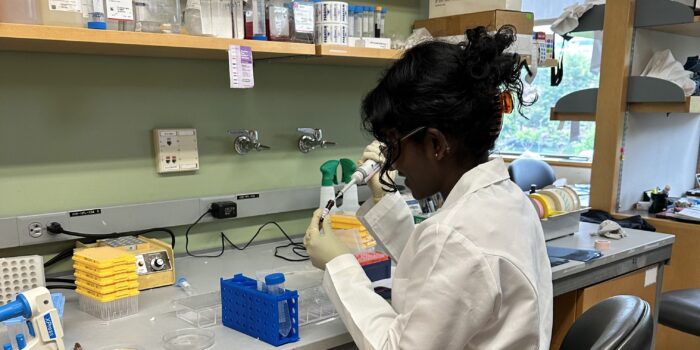 Undergraduate medic Kujani Wanniarachchi at Yale University. Here she is in a white lab coat holding a pipette in a laboratory.