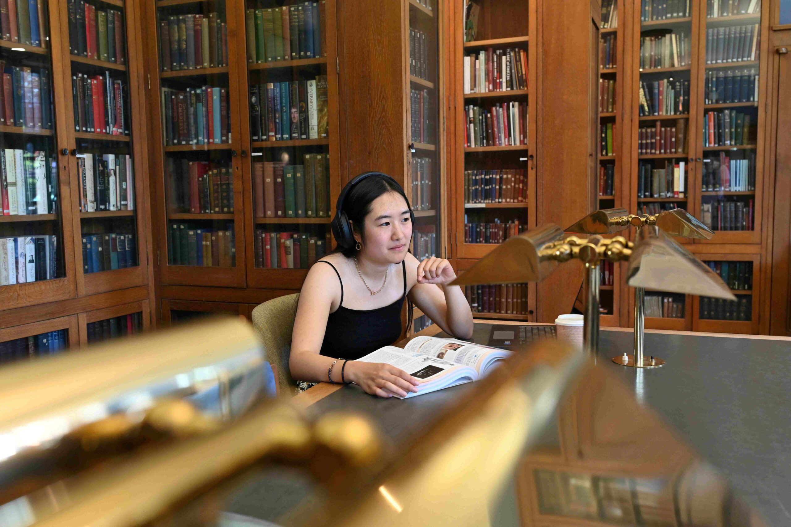 A student sits at a desk in a library with brass lamp in the foreground