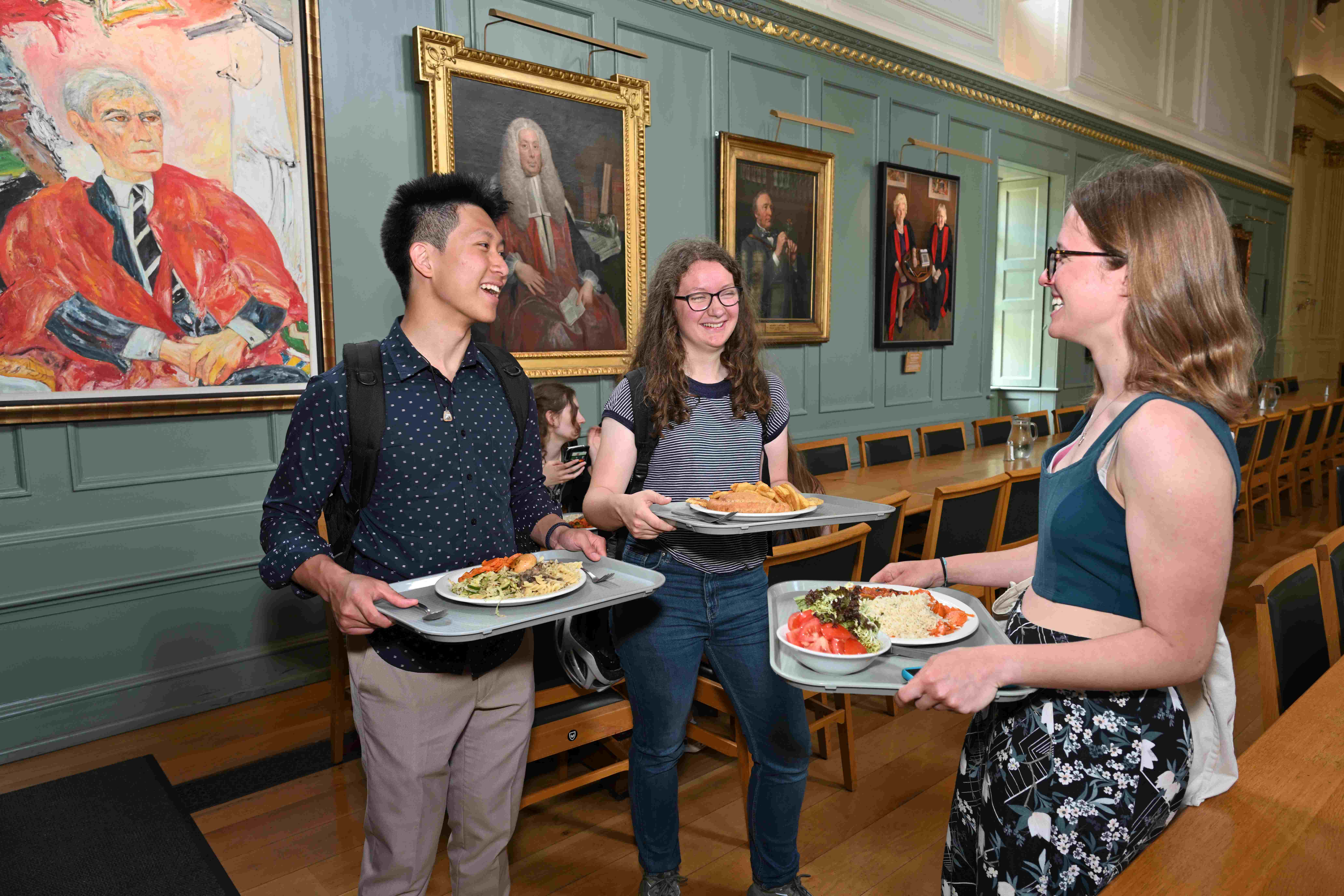 People smiling holding their food on trays in the dining hall at Trinity Hall, Cambridge.