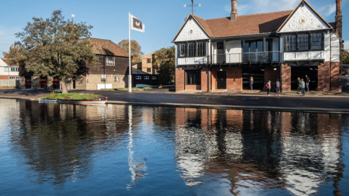 Trinity Hall boathouse next to the River Cam. The sky is blue and the College flag is flying outside