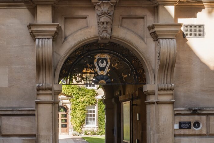 Front entrance to Trinity Hall: a large opening in a stone building with a black and white crest above and a stone face carved into the keystone which has a stone-carved seashell above it
