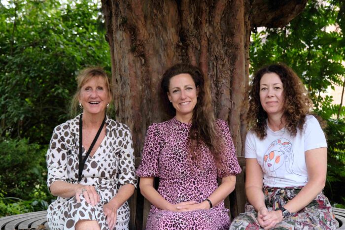 Three smiling women sitting beneath a tree. They are the Trinity Hall (Cambridge) Wellbeing Team.