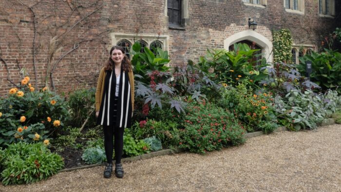 A woman, Rhiannon Warren, stands outside an old building (the Old Library) in Trinity Hall, Cambridge. The building is surrounded by plants in a beautiful flower bed.