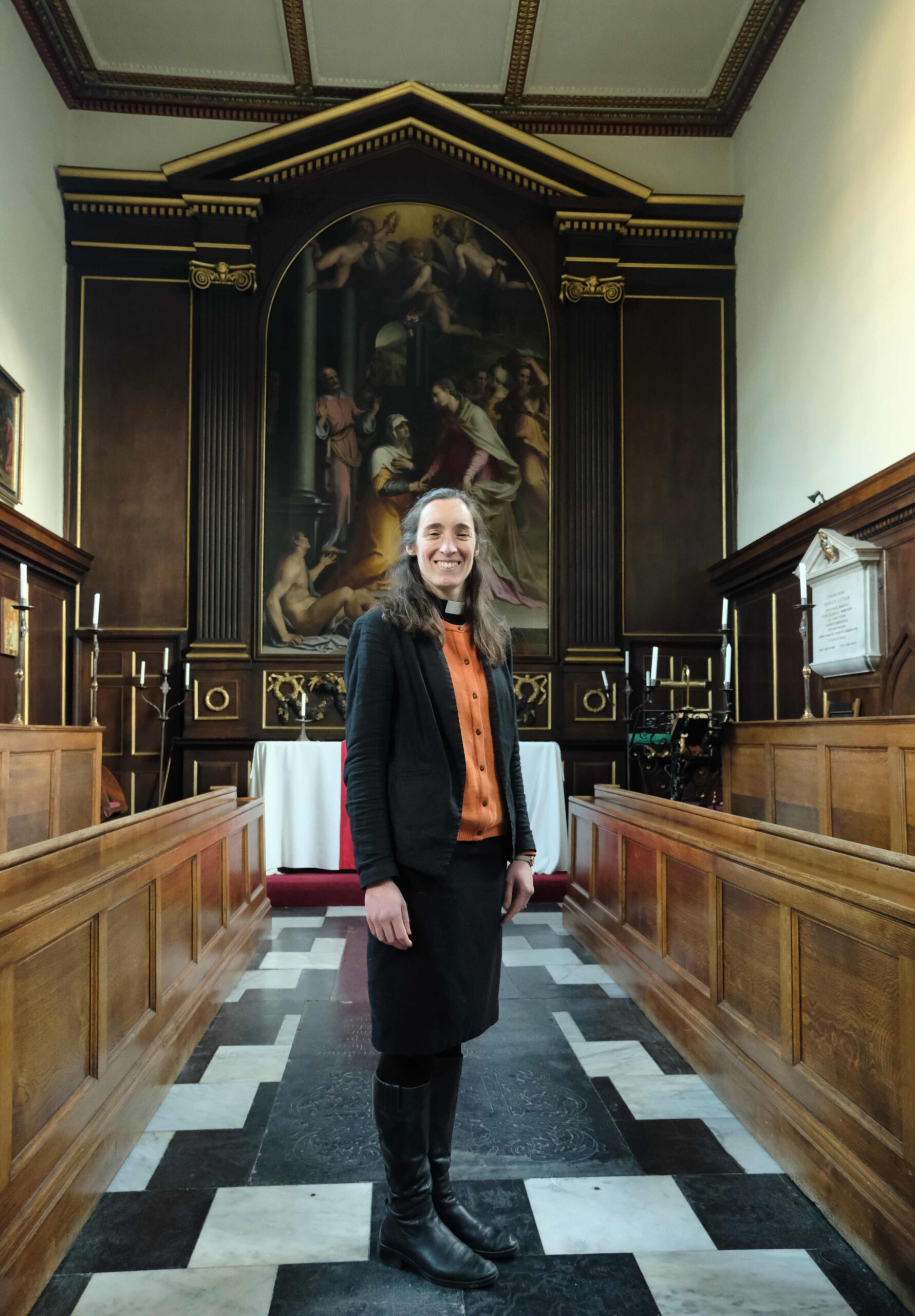The Reverend Jennifer Totney stood inside Trinity Hall Chapel. The floor is black and white tiles and behind her is a large religious painting behind an altar. She is Acting Dean at the College.
