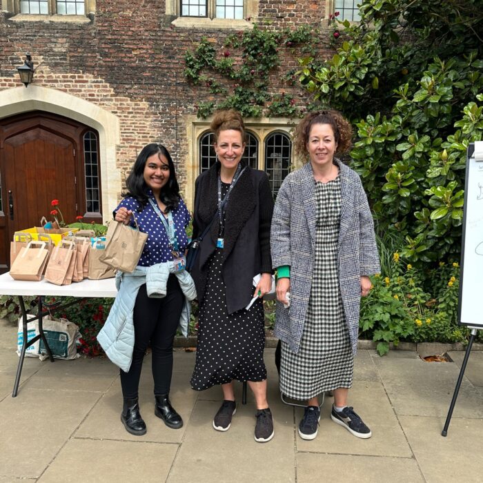 Three staff members standing in front a table of paper bags filled with exam supplies, handing them out to students in front of a historic building.
