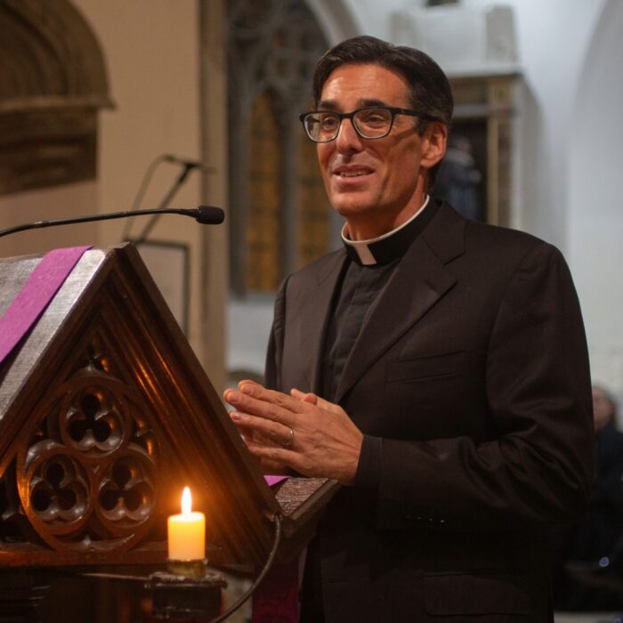 The image shows a man dressed in black stood by a dais with a Christmas tree in the background. He is The Revd Dr Mark Scarlata, Vicar-Chaplain of St Edward’s and he is introducing a talk about Robert Barnes' revolutionary sermon that sparked the Reformation in England