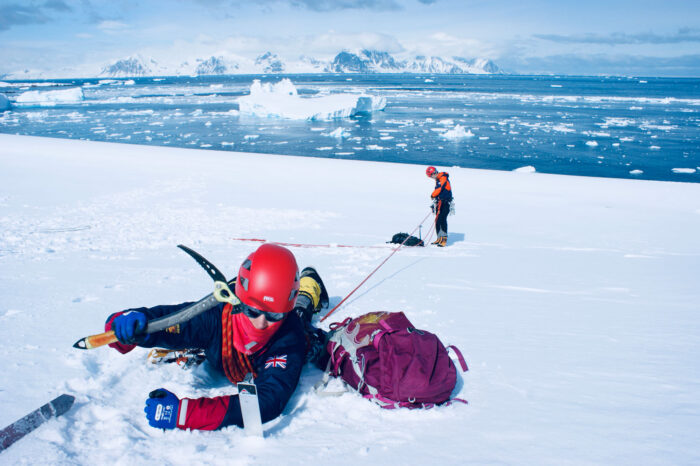 Person practicing crevasse rescue on a snowy glacier, secured by ropes, with another climber and ice-filled sea in the background.