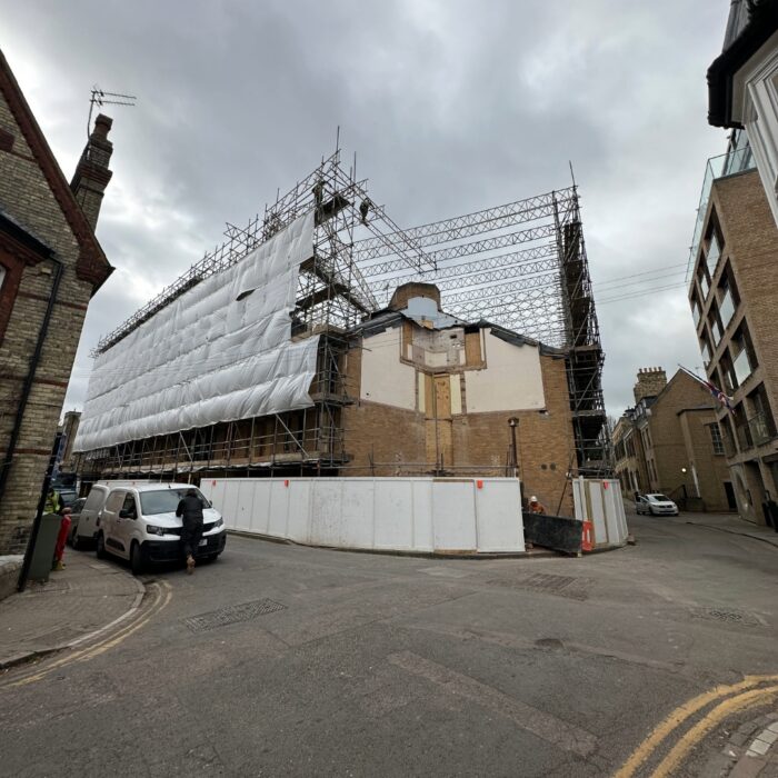 A street‑level photograph of a building partially covered in scaffolding and white protective sheeting during construction work. Temporary barriers surround the site, with neighbouring buildings and parked cars visible along the street.