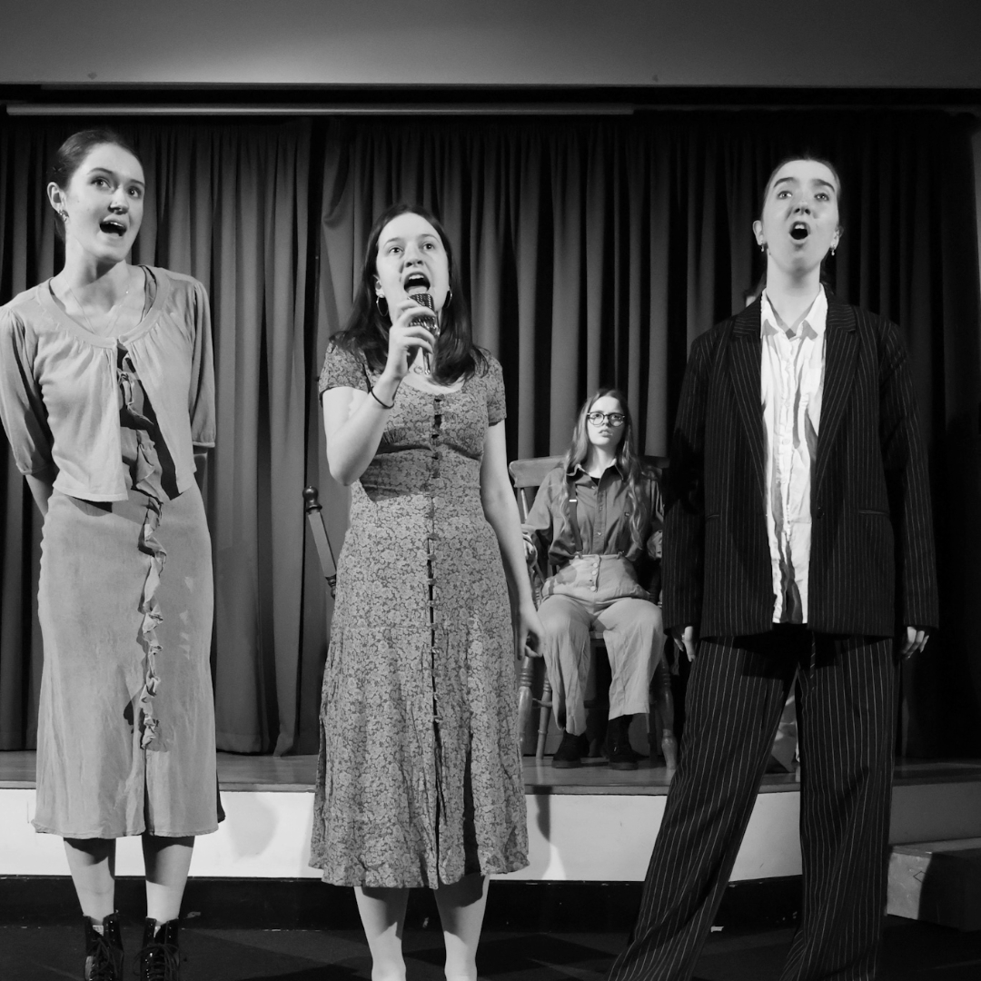 A black‑and‑white photograph of four performers standing on a stage. Three performers sing centre stage while a fourth performer sits behind them on a chair. All are wearing period‑style costumes as part of a stage scene.