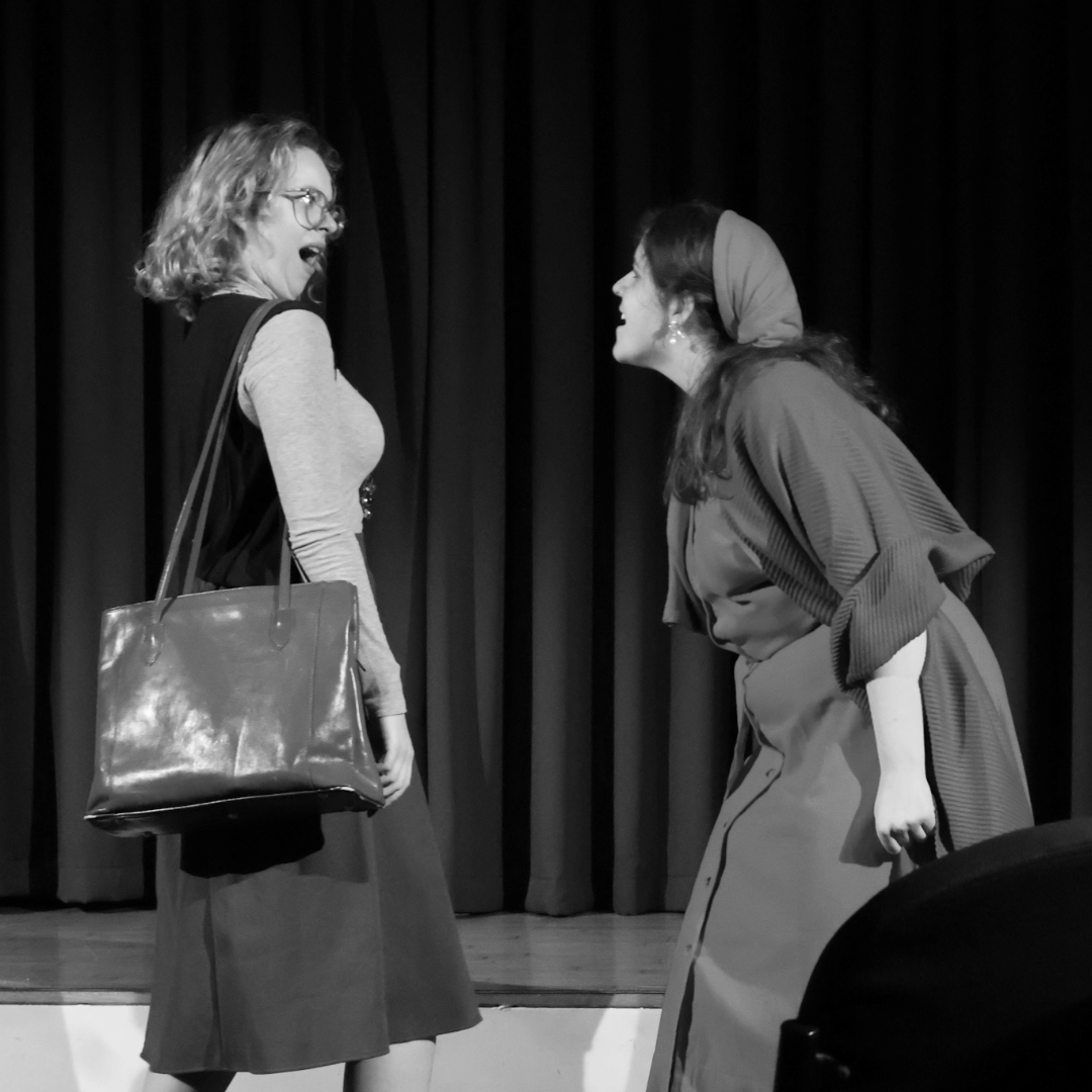 A black‑and‑white photograph of two performers on stage in front of a curtain. Both are wearing period‑style costumes. One performer stands facing the other with a large bag over one shoulder, while the second performer leans forward and gestures during the scene.