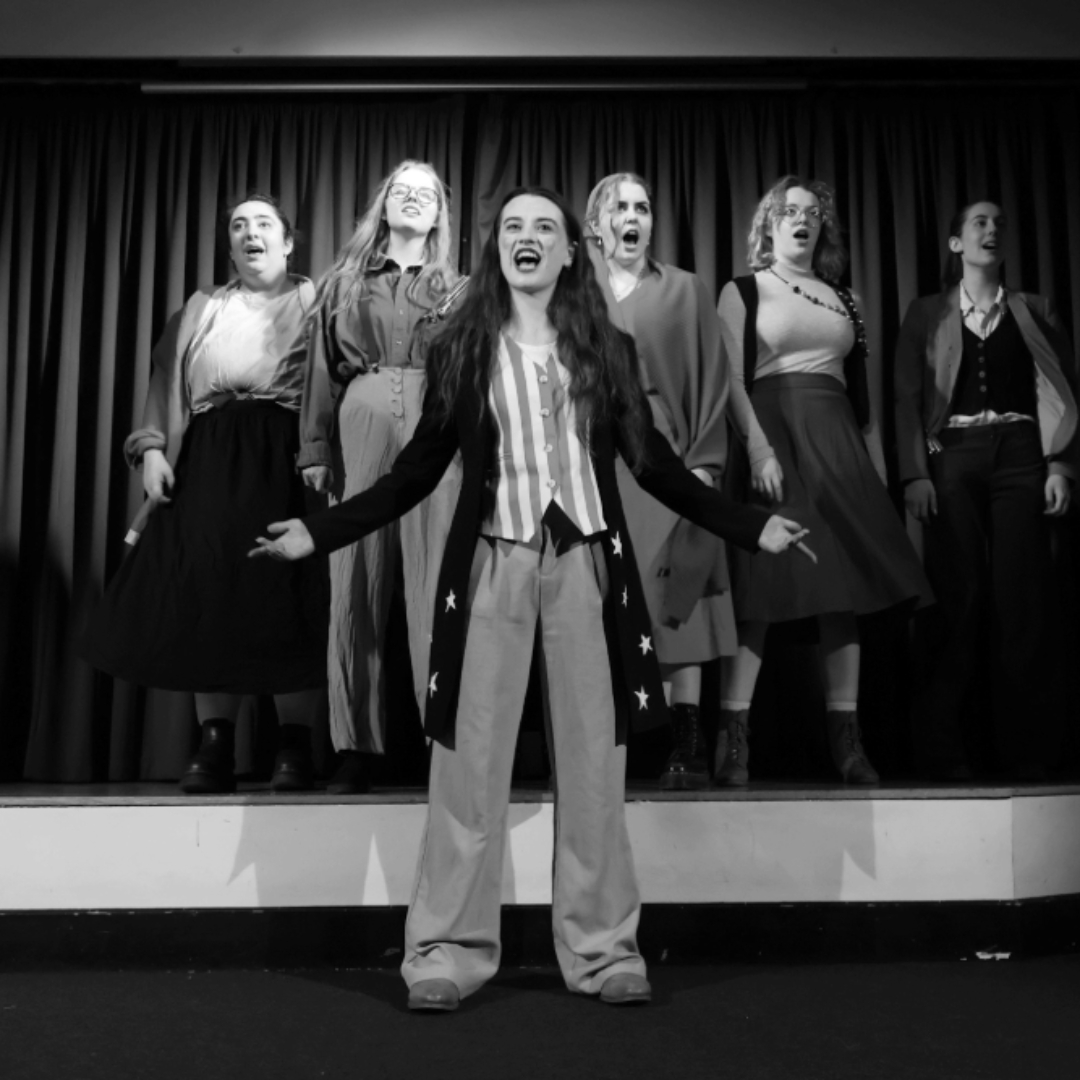 A black‑and‑white photograph of a group of performers standing on a stage in front of a curtain. One performer stands at the front, with the rest of the group positioned behind them, all wearing period‑style costumes and arranged as part of a stage scene.
