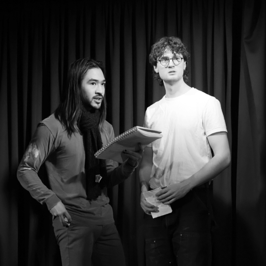 A black-and-white photograph of two performers standing on a stage in front of black curtains, with one holding a notebook and the other holding paper, appearing to rehearse or read lines under stage lighting.