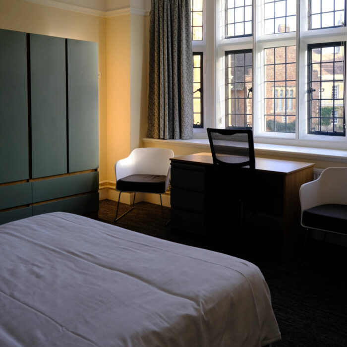 A photograph of a furnished bedroom with a neatly made bed in the foreground. A desk and chair sit by large multi‑pane windows, with upholstered chairs on either side. Built‑in storage cupboards line one wall, and natural light fills the room.