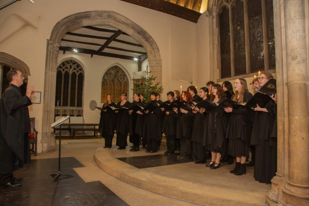 A choir standing on a raised area inside a chapel, singing from open music folders while a conductor stands at a music stand in front of them.