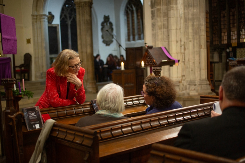 People seated in wooden pews inside a chapel, with one person leaning forward to speak to others during an event.