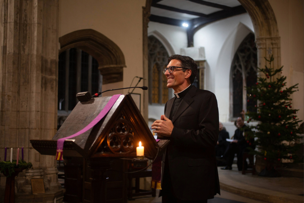 A speaker standing at a lectern inside a chapel, addressing an audience, with a lit candle and a decorated tree visible in the background.