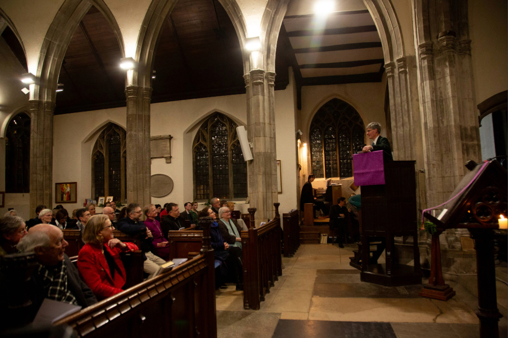 A wide view of a chapel interior showing an audience seated in pews and a speaker standing at a pulpit beneath arched stone architecture.
