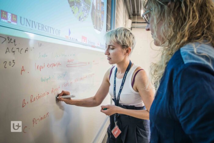 A photograph of two people standing beside a whiteboard in a classroom or workshop setting. One person is writing on the whiteboard with a marker while the other stands nearby, looking on. Notes and diagrams are visible on the board.