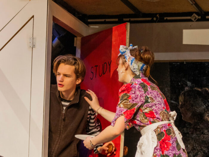 Two performers on a stage set, with one standing in a doorway speaking to another dressed in patterned clothing, with stage props and theatrical lighting visible.