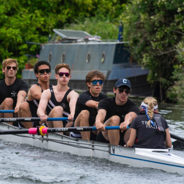 A rowing crew wearing sunglasses, powering down the river with a narrowboat in the background.