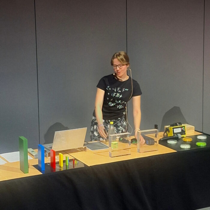 Person wearing a headset presents a science demonstration behind a table with colourful blocks, equipment and a laptop in a lecture hall.