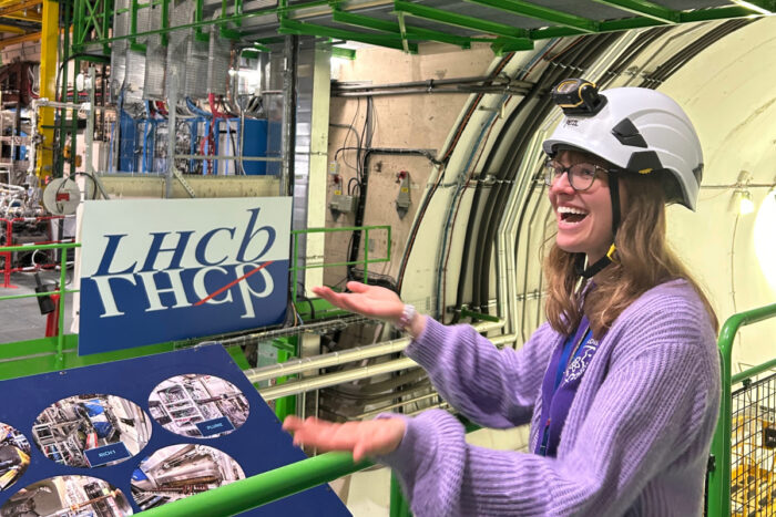 Person wearing a safety helmet stands inside the CERN Large Hadron Collider, gesturing beside equipment marked “LHCb”.