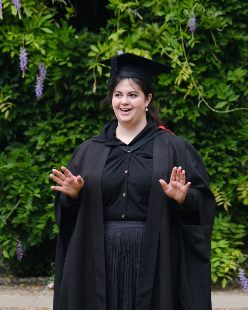 The Praelector in academic dress standing in front of leafy greenery and flowering vines.