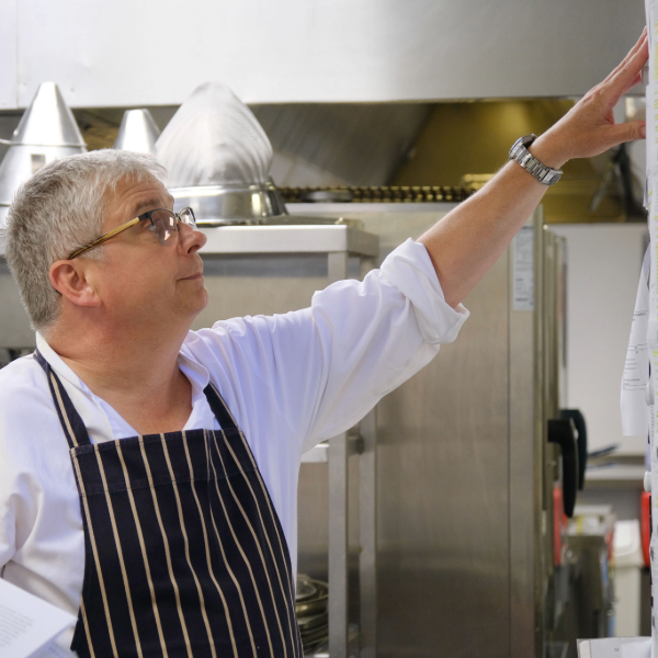 Person wearing a striped apron stands in a commercial kitchen, reaching towards a notice board beside stainless steel catering equipment.