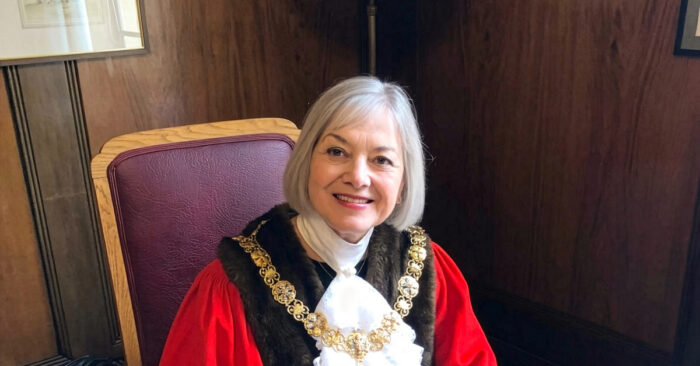 Person wearing ceremonial robes and a gold chain sits in a high-backed chair in a wood-panelled room.