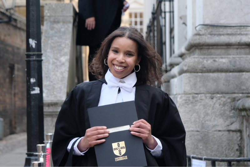 Student in formal attire walking down the steps of a historic building, holding a degree certificate.