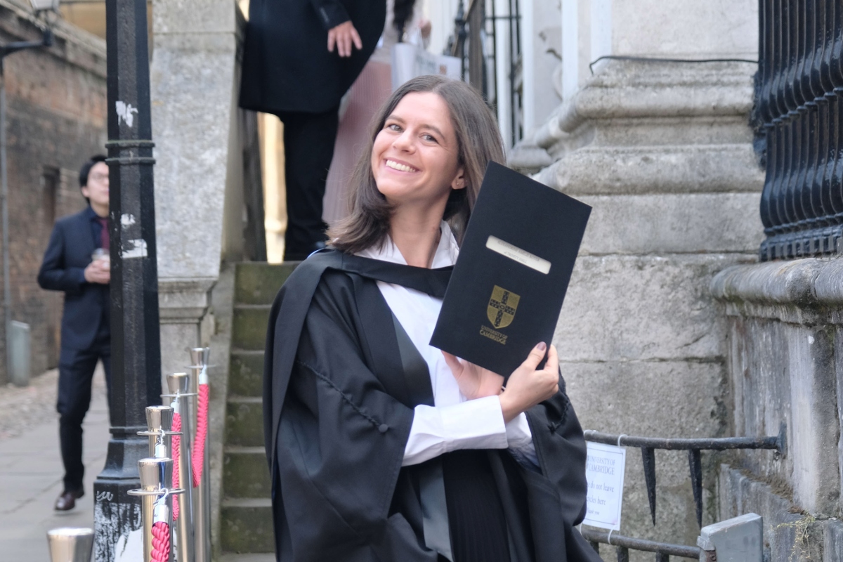 Person in academic dress stands outside a historic stone building, holding a University of Cambridge certificate folder.