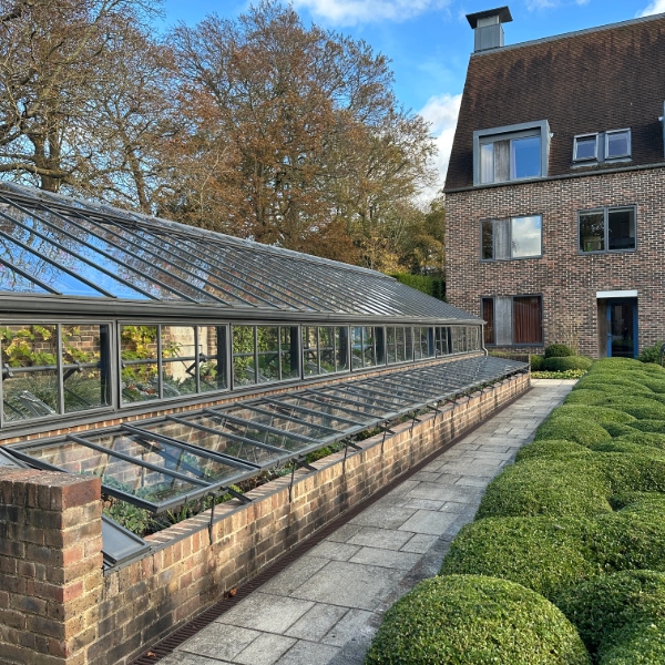 Glass-roofed greenhouse beside a brick building, with a paved walkway, clipped hedges, and trees under a blue sky.