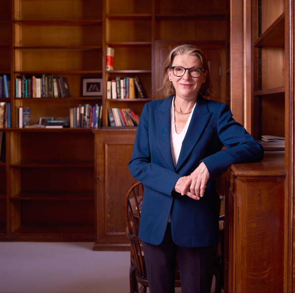Person wearing glasses and a blue jacket stands in a wood-panelled library, leaning against a bookcase with shelves of books in the background.