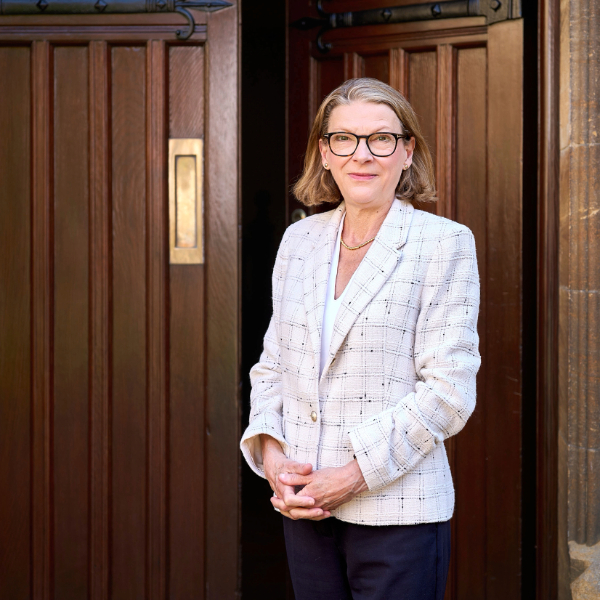 Portrait of a person in a light checked blazer standing in a wooden doorway.