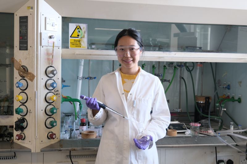A person in a white lab coat standing amongst scientific equipment, holding a pipette and vesicle.