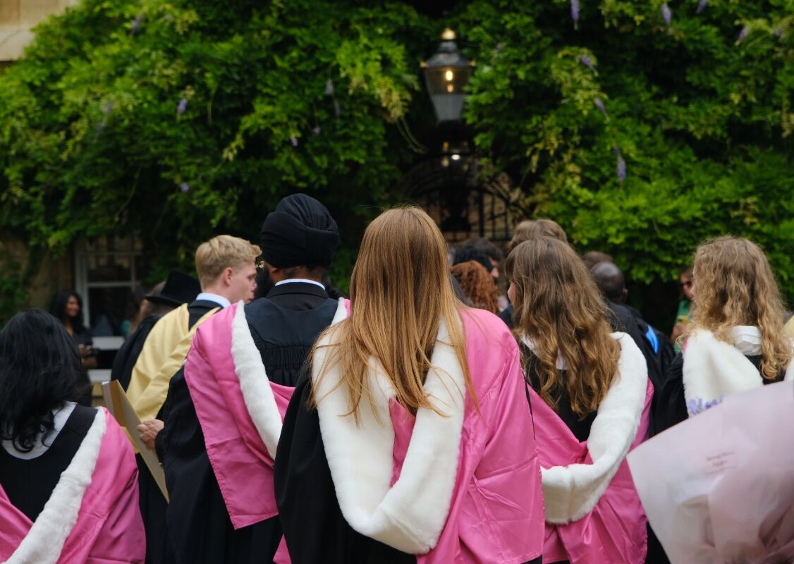 Graduates wearing pink and white academic hoods gather beneath a leafy archway covered in green foliage.