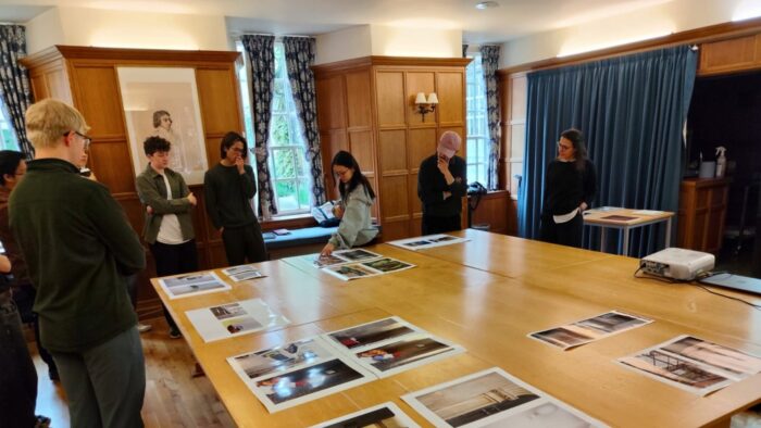 A group of people standing around a large wooden table in a wood-panelled room, discussing and examining printed photographs laid out on the table.