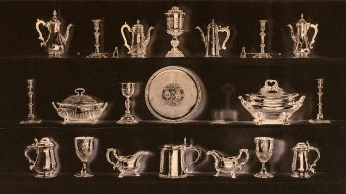 Historic display of silver tableware arranged on four shelves, including cups, candelabras, teapots, candlesticks, serving dishes and plates, shown in a sepia-toned photograph.