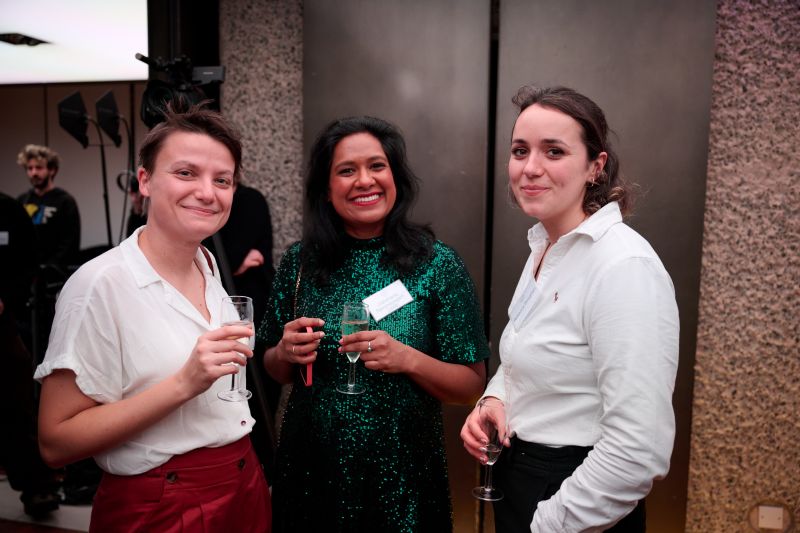 Three alumni holding drinks, smiling, at the 675th Birthday Party at the Barbican.