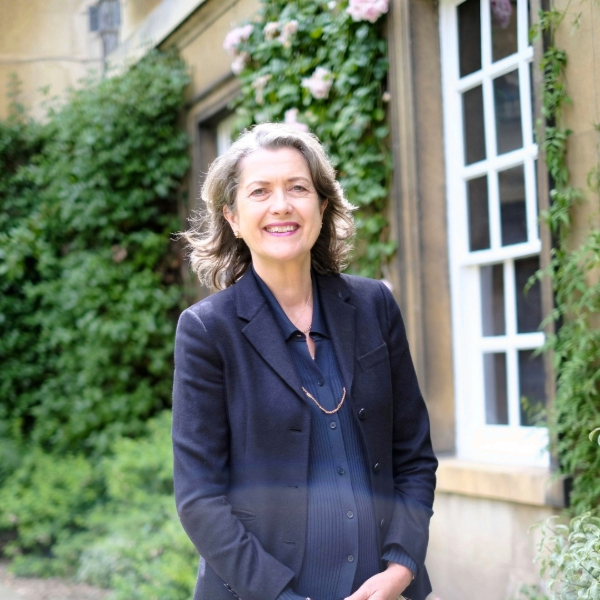 Person in a dark suit stands in a garden beside a vine-covered historic building, with a window and flowering greenery in the background.
