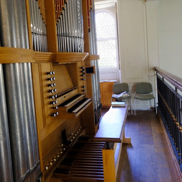 Organ console with keyboards, pedals and rows of stops inside a chapel gallery, beside a glass window.