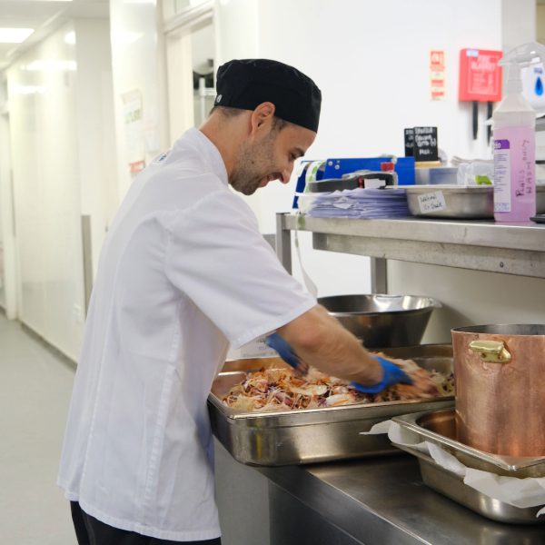 Kitchen staff member preparing food in a commercial kitchen, mixing ingredients in a large metal tray beside cooking equipment and utensils.